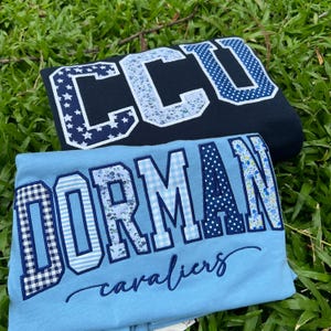 May include: A navy blue sweatshirt with the letters CCU in white-bordered patterned fabric, and a light blue t-shirt featuring the word DORMAN in varied blue patterned fabric with the word cavaliers embroidered in dark blue script below.