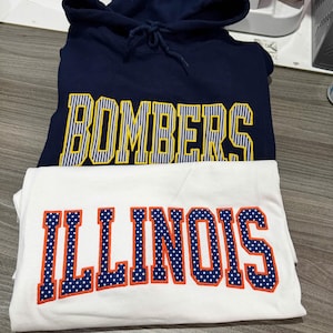 May include: A navy blue hooded sweatshirt with the word BOMBERS in yellow and white striped lettering, layered over a white t-shirt featuring the word ILLINOIS in blue and white polka-dot lettering with a bright orange outline.
