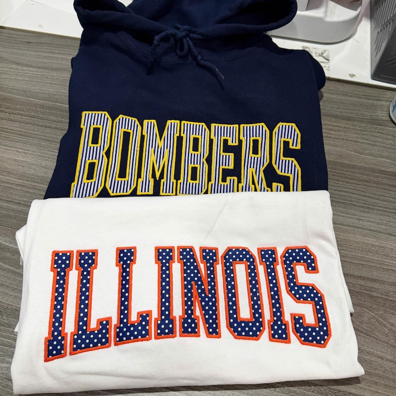 May include: A navy blue hooded sweatshirt with the word BOMBERS in yellow and white striped lettering, layered over a white t-shirt featuring the word ILLINOIS in blue and white polka-dot lettering with a bright orange outline.