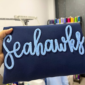 May include: A dark navy blue sweatshirt featuring the word Seahawks embroidered in a light blue, textured, cursive script font across the front. The lettering has a raised, fuzzy appearance that stands out against the smooth fabric surface.