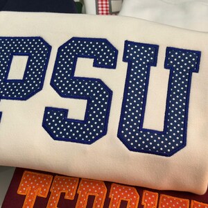 May include: A cream-colored sweatshirt featuring the letters PSU in large, block-style font. The letters are dark blue with a white polka dot pattern and are outlined in dark blue embroidery, creating a bold, textured appearance on the fabric.
