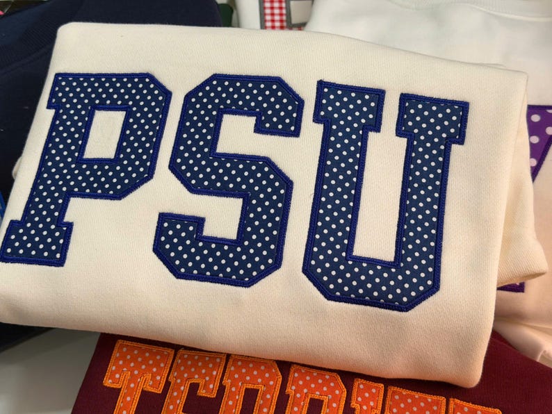 May include: A cream-colored sweatshirt featuring the letters PSU in large, block-style font. The letters are dark blue with a white polka dot pattern and are outlined in dark blue embroidery, creating a bold, textured appearance on the fabric.