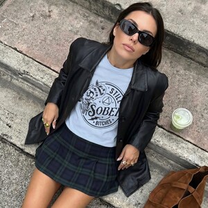 May include: A light blue t-shirt with the text "STILL SOBER BITCHES" is worn under a black leather jacket. A green and blue plaid skirt and black knee-high boots complete the outfit. A brown suede tote bag and sunglasses are also visible.