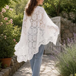 May include: A white crochet poncho featuring an intricate circular floral pattern throughout the fabric, with a scalloped edge along the hemline. The garment is worn over light blue denim jeans, creating a layered look with a textured finish.