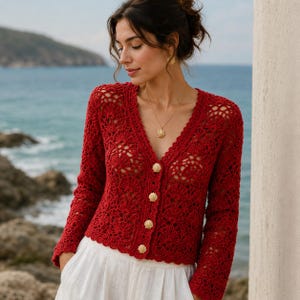 May include: A long-sleeved, deep red crochet cardigan featuring an intricate openwork shell pattern. The garment has a V-neckline and is fastened down the front with five gold-toned buttons shaped like seashells. The knit texture is consistent throughout.