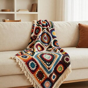 May include: A colorful, crocheted blanket with a geometric pattern of squares and circles in shades of blue, orange, red, and green. The blanket has a cream-colored fringe along the edges and is draped over a beige sofa.
