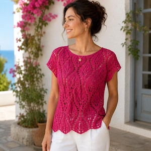 May include: A magenta-colored, short-sleeved top with an openwork leaf pattern. The top has a round neckline and a scalloped hem. The woman is wearing white pants. The background includes pink flowers and a view of the ocean.