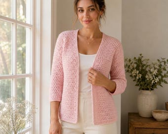 May include: A light pink open-front cardigan featuring a textured crochet knit pattern, three-quarter length sleeves, and a straight hemline. The garment is worn over a simple white top and light-coloured trousers, showcasing a clean, layered look.