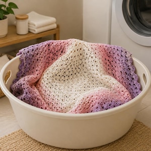 May include: A crochet blanket featuring an ombre color gradient that transitions from a deep purple at the edges to a soft pink and finally to a creamy white center. The blanket has an intricate, open-work stitch pattern and a scalloped border.