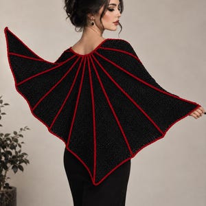 May include: A black crocheted capelet featuring a bat wing design with prominent red trim along the structural ribs and edges. The garment is worn over a black top, showcasing a wide, pointed silhouette that extends outward from the shoulders.