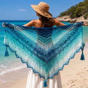 May include: A triangular crochet shawl featuring an ombre gradient pattern that transitions from deep navy blue at the center to bright turquoise and white at the edges, finished with three matching blue tassels at the corners and points.