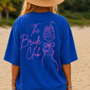 Op de afbeelding: Een koningsblauw t-shirt met een roze afbeelding op de achterkant. Het ontwerp bevat de tekst "The Bride Club" in een cursief lettertype naast een lijntekening van een cocktailglas met een strik, "18. JULI 2026" en "MELLI'S JGA".