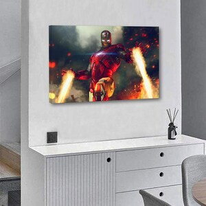 May include: A canvas print featuring Iron Man in a dynamic pose, with his hands emitting bright energy beams. The artwork showcases the superhero in his iconic red and gold armor against a fiery, smoky background. The print is mounted on a white wall.