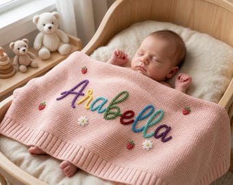 Könnte beinhalten: Ein schlafendes Baby liegt in einer Holzwiege und ist mit einer rosa Strickdecke bedeckt. Auf der Decke ist der Name "Arabella" in bunten Fäden sowie Erdbeer- und Gänseblümchen-Akzente gestickt. Ein Teddybär und andere Spielsachen sind in der Nähe zu sehen.