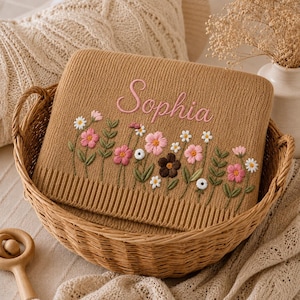 May include: A tan knitted blanket with the name "Sophia" embroidered in pink script. The blanket features a floral design with pink, white, and brown flowers, and green leaves. The blanket is in a woven basket.