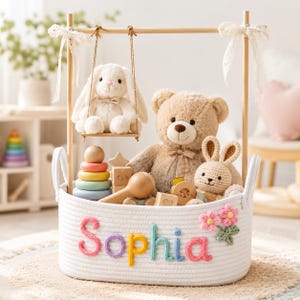 Puede incluir: Una cesta de cuerda blanca llena de juguetes, incluyendo un oso de peluche, un conejito de ganchillo y bloques de madera. Un conejito de peluche está sentado en un columpio encima de la cesta. El nombre "Sophia" está bordado al frente en letras coloridas.