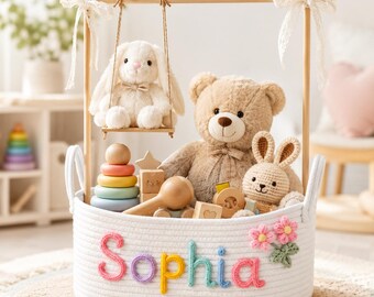 Könnte beinhalten: Ein weißer Seilkorb gefüllt mit Spielzeug, darunter ein Teddybär, ein gehäkelter Hase und Holzbausteine. Ein Plüschhase sitzt auf einer Schaukel über dem Korb. Der Name "Sophia" ist in bunten Buchstaben auf die Vorderseite gestickt.