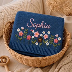 May include: A navy blue knitted blanket with the name "Sophia" embroidered in pink script. The blanket features a floral design with pink, white, and brown flowers and green leaves. The blanket is folded in a wicker basket.