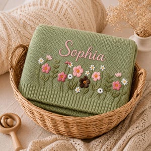May include: A sage green knitted blanket with the name "Sophia" embroidered in pink script. The blanket features a floral design with pink, white, and brown flowers. It is displayed in a woven basket.