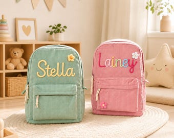 Könnte beinhalten: Zwei Rucksäcke aus Cord, einer in Salbeigrün mit dem Namen Stella und einer gelben Blume, und einer in Altrosa mit dem Namen Lainey in bunten Buchstaben und zwei kleinen Blumen. Beide Taschen haben eine Fronttasche mit Reißverschluss und weiße Kordeln.