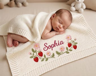 Könnte beinhalten: Ein schlafendes Baby liegt auf einer cremefarbenen Strickdecke. Die Decke ist mit dem Namen "Sophia", rosa Blumen und roten Erdbeeren bestickt. Das Baby ist in eine passende cremefarbene Decke gehüllt.