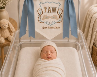Könnte beinhalten: Ein neugeborenes Baby, eingewickelt in eine cremefarbene Decke und Mütze, liegt in einer transparenten Babywanne. Darüber ein Banner mit den Initialen "TAW" und dem Namen "Tyler Wells Abke", bestickt mit Cowboyhut und Hufeisen, und zwei blauen Schleifen.
