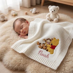Op de afbeelding: Een slapende baby ligt op een crèmekleurig, zacht tapijt, bedekt met een witte gebreide deken. De deken heeft een geborduurd beeld van Winnie de Poeh en Teigetje, met de naam "Noah" in roze script.