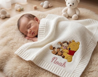 Könnte beinhalten: Ein schlafendes Baby liegt auf einem cremefarbenen, flauschigen Teppich, bedeckt von einer weißen Strickdecke. Die Decke zeigt ein gesticktes Bild von Winnie Puuh und Tigger mit dem Namen "Noah" in rosa Schrift.