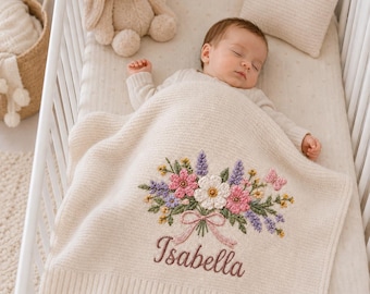 Könnte beinhalten: Eine cremefarbene Strickdecke für Babys mit einem gestickten Blumenstrauß in Rosa, Weiß und Violett, einem kleinen rosa Schmetterling und einer rosa Schleife. Der Name Isabella ist darunter in brauner Schreibschrift eingestickt.