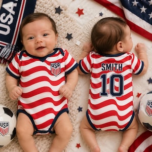 Pode incluir: Dois bodies de manga curta para bebé com um padrão de riscas onduladas vermelhas e brancas e acabamentos em azul escuro. A frente apresenta um emblema de escudo USA. As costas exibem o nome SMITH e o número 10 em letras azul escuro.