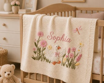 Könnte beinhalten: Cremefarbene Babydecke mit gesticktem Blumen- und Schwanenmuster. Die Decke zeigt den Namen "Sophia" in rosa Schrift, zusammen mit rosa und gelben Blumen, einer Libelle und einem weißen Schwan mit orangem Schnabel.