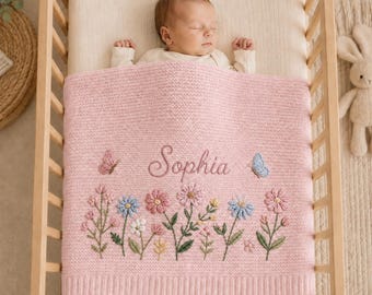 Könnte beinhalten: Eine weiche, rosa gestrickte Babydecke mit dem eingestickten Namen Sophia in Schreibschrift, verziert mit bunten Blumenstielen und zwei kleinen Schmetterlingen. Die Decke hat ein strukturiertes Strickmuster und einen gerippten Rand.
