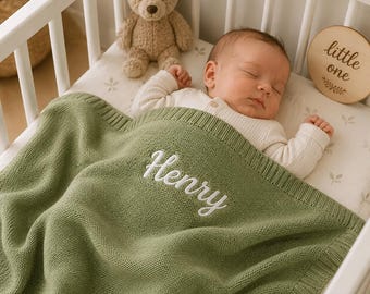 Könnte beinhalten: Eine salbeigrüne Strickdecke für Babys mit dem in weißer Schreibschrift eingestickten Namen Henry. Die Decke hat einen gerippten Rand und liegt über einem schlafenden Säugling, der ein cremefarbenes Langarmshirt in einem weißen Kinderbett trägt.