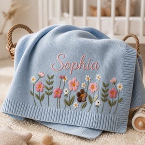 May include: A light blue baby blanket with embroidered flowers in pink, white, and brown. The name "Sophia" is embroidered in pink script. The blanket is folded in a wicker basket.