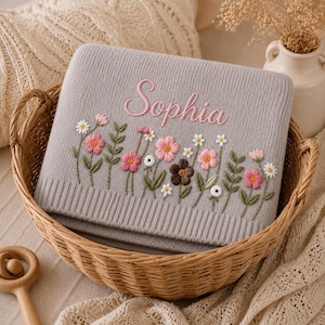May include: A gray baby blanket embroidered with the name "Sophia" in pink script. The blanket features a floral design with pink, white, and brown flowers, and green leaves. The blanket is folded in a wicker basket.