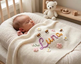 Könnte beinhalten: Ein schlafendes Baby liegt in einer weichen, cremefarbenen Babywiege, bedeckt mit einer cremefarbenen Strickdecke. Die Decke ist mit gestickten Blumen, Bienen und dem Namen "Luna" in bunten Garn verziert. Ein gehäkelter Teddybär sitzt in der Nähe.
