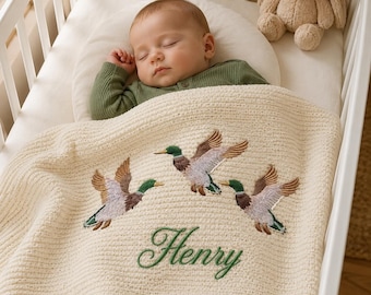 Könnte beinhalten: Eine cremefarbene Strickdecke für Babys mit einer Stickerei von drei fliegenden Stockenten in Grün-, Braun- und Weißtönen, wobei der Name Henry in grüner Schreibschrift unter den Enten auf dem strukturierten Stoff eingestickt ist.