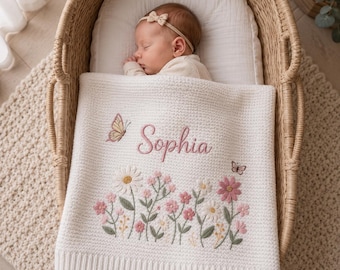 Könnte beinhalten: Eine weiße gestrickte Babydecke, bestickt mit dem Namen "Sophia" in rosa Schrift, sowie einem Blumendesign mit Gänseblümchen und rosa Blumen. Zwei Schmetterlinge sind ebenfalls auf die Decke gestickt. Die Decke liegt in einer Wiege aus Korbgeflecht.
