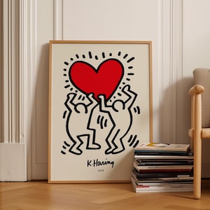 May include: A framed art print featuring a bold black line drawing of two stylised figures holding up a large, solid red heart. The background is a cream colour. Below the figures, the text reads K. Haring and the word LOVE in black lettering.