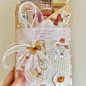 May include: A brown paper package labelled Blind date with a book Romance Edition, decorated with a white organza ribbon, a gold paperclip, butterfly accents, and floral stickers. The package contains a book review card, library card, and snacks.