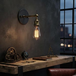 Pode incluir: Um candeeiro de parede com base circular preta e braço de latão que sustenta uma lâmpada Edison de vidro transparente. O candeeiro está posicionado sobre uma secretária de madeira com engrenagens de metal, um pequeno relógio preto e um caderno.