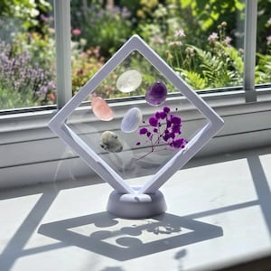 May include: A white diamond-shaped floating display frame containing five smooth polished gemstones in shades of clear, purple, pink, blue, and speckled grey, alongside a small sprig of dried purple flowers, all mounted on a circular white base.