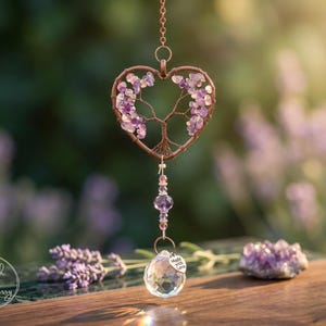 May include: A heart-shaped suncatcher featuring a copper wire tree of life design with purple amethyst chip leaves. A vertical strand of amethyst beads and a clear faceted crystal prism hangs below, accented by a small tag reading Made with Love.