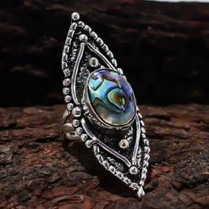 May include: A silver ring featuring an elongated, marquise-shaped setting with intricate metalwork and beaded edges. The centre holds an oval, iridescent abalone shell inlay displaying swirling patterns of blue, green, and purple hues.