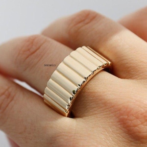 Peut inclure: Une bague dorée composée d'un large anneau plat orné d'une série de crêtes verticales et arrondies sur toute la surface extérieure. Le mot SPIEWELS est imprimé en petites lettres noires sur le côté de l'anneau, qui présente une finition polie.