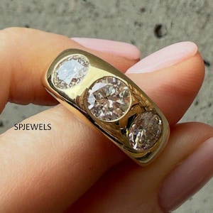 Puede incluir: Un anillo de banda dorada con tres piedras redondas, transparentes y facetadas dispuestas en fila horizontal. El metal tiene un acabado pulido y reflectante. El texto SPJEWELS está impreso en negro en el lateral del anillo cerca de la piedra.