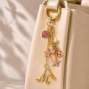 Op de afbeelding: Een crèmekleurige handtas met een goudkleurige metalen tashanger. De hanger bestaat uit een gouden ketting met meerdere bedels, waaronder een rood hart, een roze zeester, een witte parel, een kleine schelp, een rood-wit geblokt vierkant en een letter M.