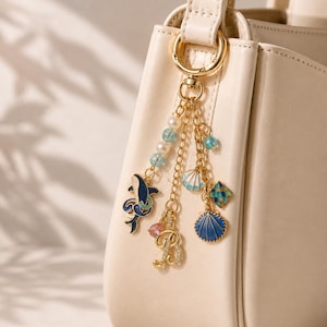 May include: A gold-toned metal bag charm attached to a cream-coloured handbag. The charm features a dangling gold chain with a blue enamel whale, a letter B, a blue and white seashell, a small blue bead, and a square blue patterned charm.