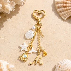 May include: A gold-toned keychain featuring a heart-shaped clasp, a dangling letter M charm encrusted with small clear stones, a white shell charm, a white starfish charm, a white whale tail charm, a small pearl, and a yellow teardrop gem.