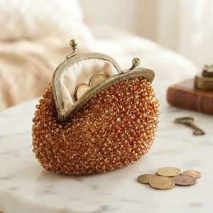 May include: A small, semi-circular coin purse covered in amber-colored glass beads with a vintage-style, antiqued brass metal frame and a kiss-lock clasp. The interior is lined with cream-colored fabric and contains several metallic coins.